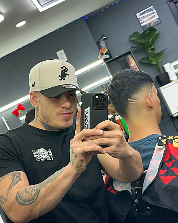 Luis, con gorra y playera negra, se toma una foto en el espejo de la barbería junto a un cliente de espaldas